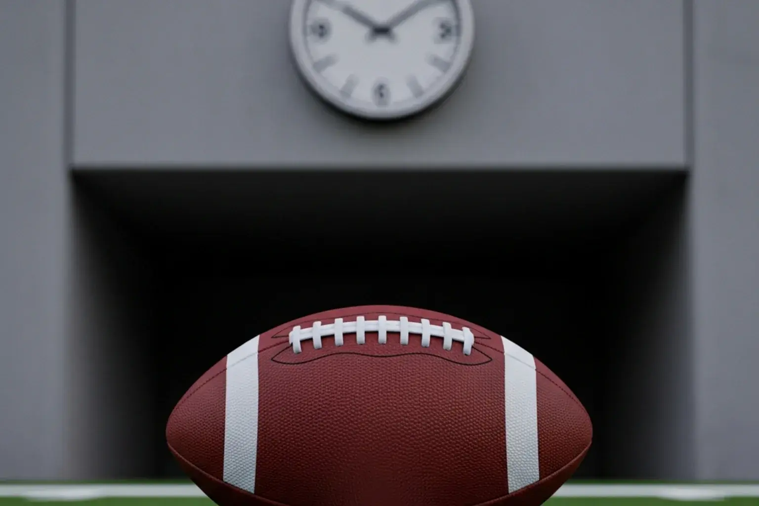 American football resting on a stadium pitch with a wall clock visible on the stadium concourse